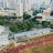 An aerial view of an urban area featuring a sports complex with a track field and a grandstand. Surrounding the sports area are numerous high-rise buildings, greenery, and roads with vehicles. The image captures people engaging in activities on the field.
