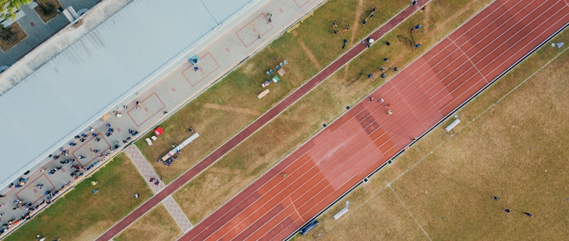 An aerial view of a sports complex featuring a running track and a grassy field. The tracks are red and situated next to a large building. There are several people scattered across the area, some on the track and others near the building. The surroundings include trees and geometric pathways, adding structure to the scene.