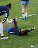 A group of athletes in sportswear are on a green soccer field. Two individuals are interacting closely; one helps the other who is lying on the grass. Nearby, other athletes are stretching.