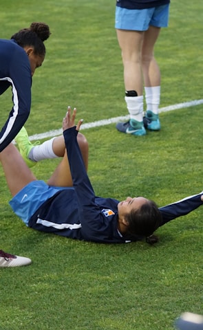 A group of athletes in sportswear are on a green soccer field. Two individuals are interacting closely; one helps the other who is lying on the grass. Nearby, other athletes are stretching.