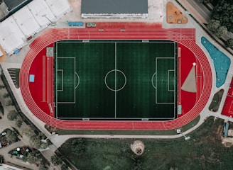 An aerial view of a large sports complex featuring a central green football field surrounded by a red running track. Adjacent to the track, there is a parking area, buildings, and pathways. Nearby, there are additional sports courts in different colors, including a blue swimming pool and orange basketball courts.