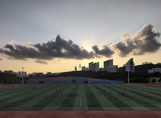 A sports field is situated in the foreground, with several people engaged in activities. The field is surrounded by a running track, and there are goalposts on either end. Behind the field, a line of modern buildings is visible, partially covering a hillside. Above, a dramatic cloud formation stretches across the sky, with the sun just below the horizon creating a silhouette effect.