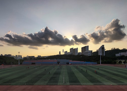 A sports field is situated in the foreground, with several people engaged in activities. The field is surrounded by a running track, and there are goalposts on either end. Behind the field, a line of modern buildings is visible, partially covering a hillside. Above, a dramatic cloud formation stretches across the sky, with the sun just below the horizon creating a silhouette effect.