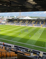A sports stadium with a well-maintained grass field is depicted. Players are spread across the field, engaging in warm-up activities. The stands are partially filled with spectators, and the stadium is surrounded by urban buildings under a partly cloudy sky.