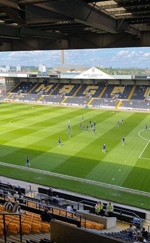 A sports stadium with a well-maintained grass field is depicted. Players are spread across the field, engaging in warm-up activities. The stands are partially filled with spectators, and the stadium is surrounded by urban buildings under a partly cloudy sky.