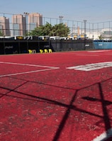 A sports court with a red playing surface and white lines, surrounded by a black mesh fence. In the background, tall buildings are visible, along with some trees and a few yellow chairs positioned near the fence.
