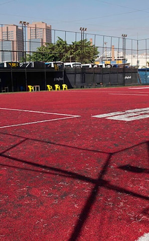 A sports court with a red playing surface and white lines, surrounded by a black mesh fence. In the background, tall buildings are visible, along with some trees and a few yellow chairs positioned near the fence.